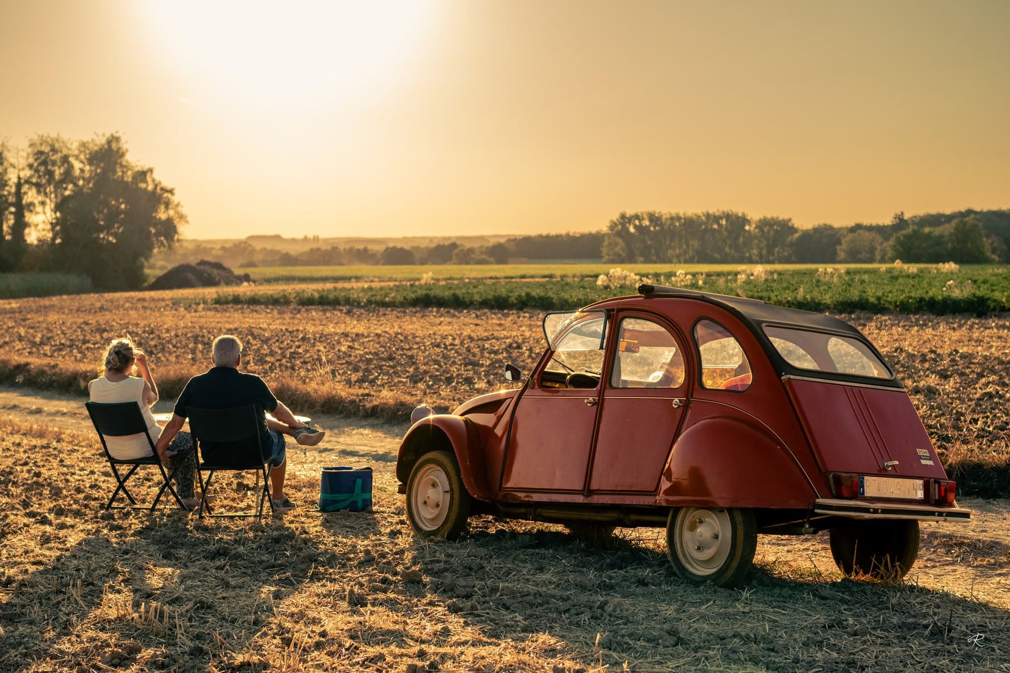 Twee mensen bij een 2CV in een stoppelveld bij zonsondergang